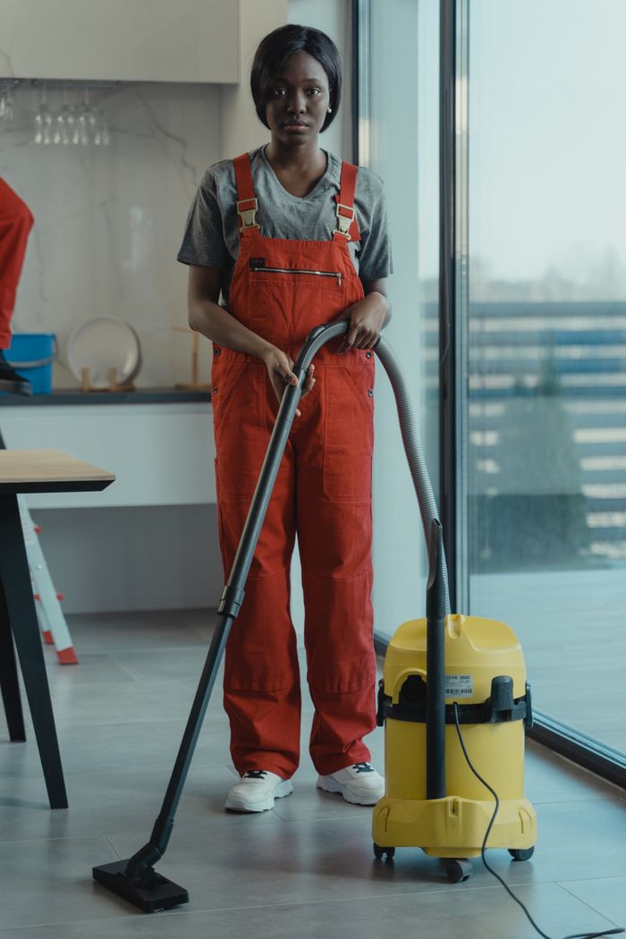 about-04 An adult woman in red coveralls operates a vacuum cleaner in a modern indoor setting, focusing on cleaning tasks.